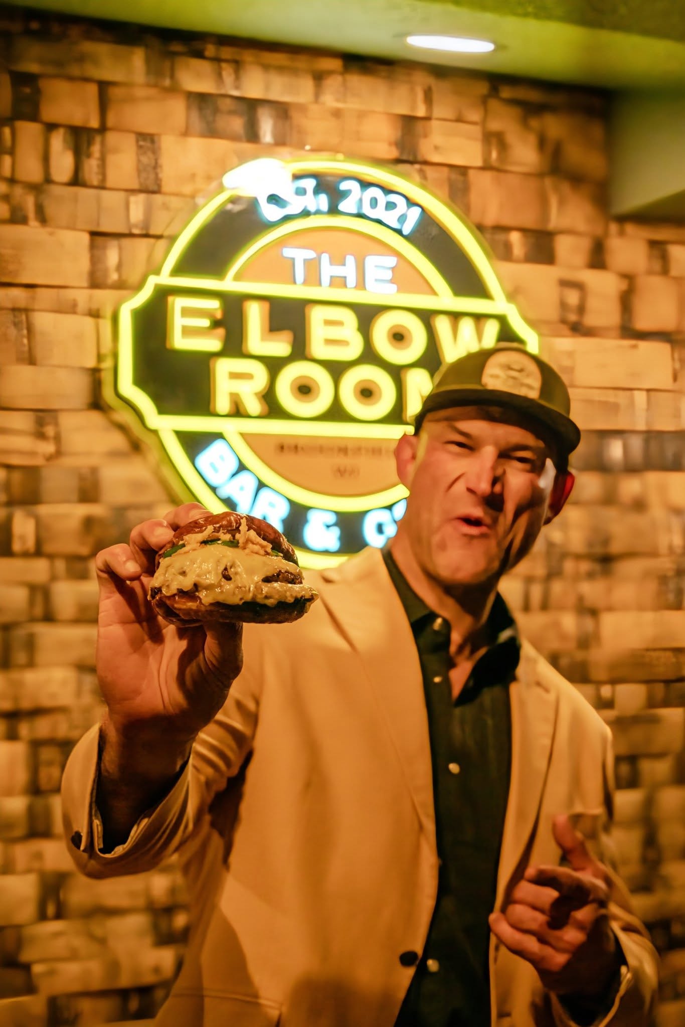 Joe holding a burger in front of 'The Elbow Room' neon sign.