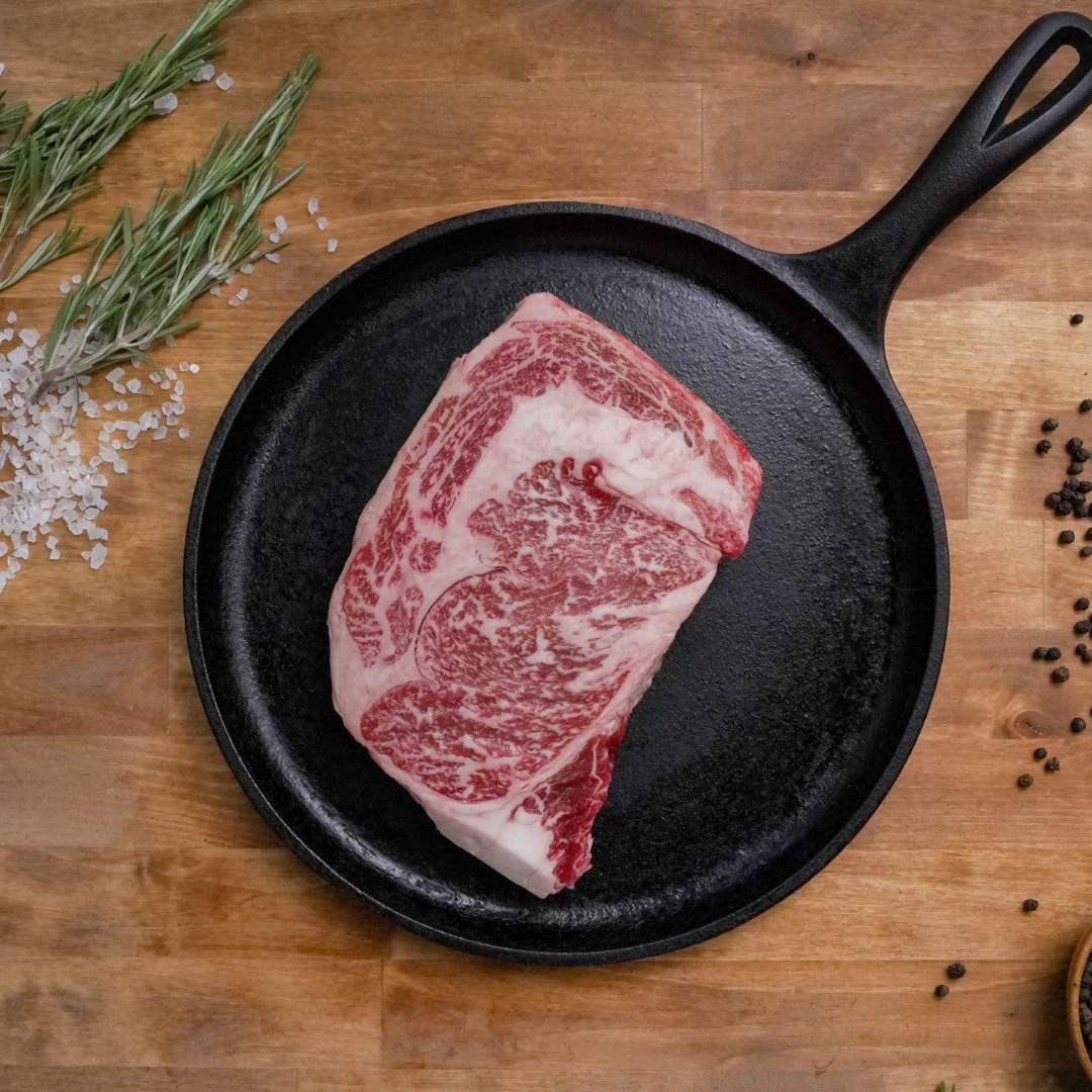 Raw Ribeye in a cast iron skillet on a wooden surface with herbs and spices.