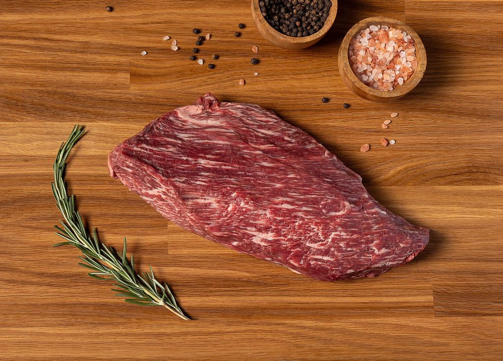 A raw marbled wagyu beef steak lies on a wooden cutting board, styled with a sprig of rosemary, scattered peppercorns, and two small wooden bowls containing black and pink salt.
